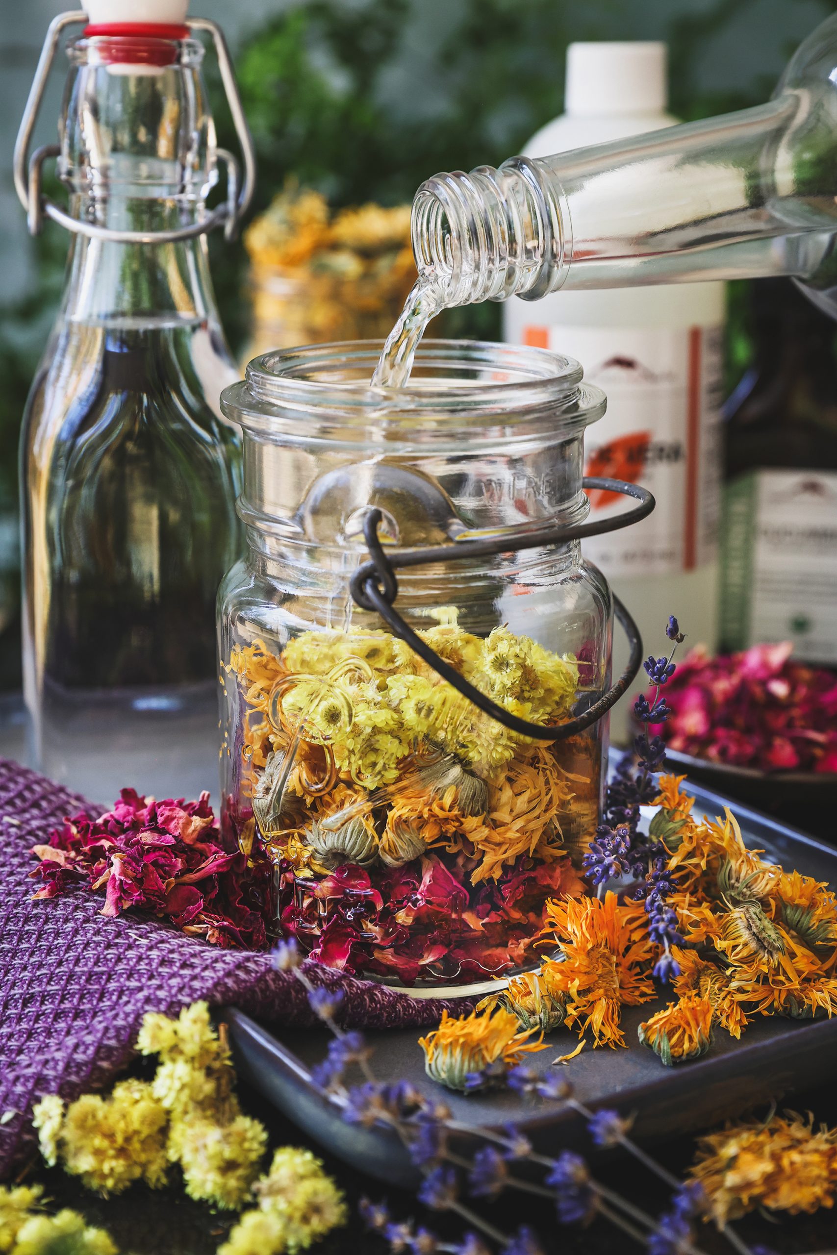 A jar of facial toner herbs with alcohol being poured into it with dried herbs around it
