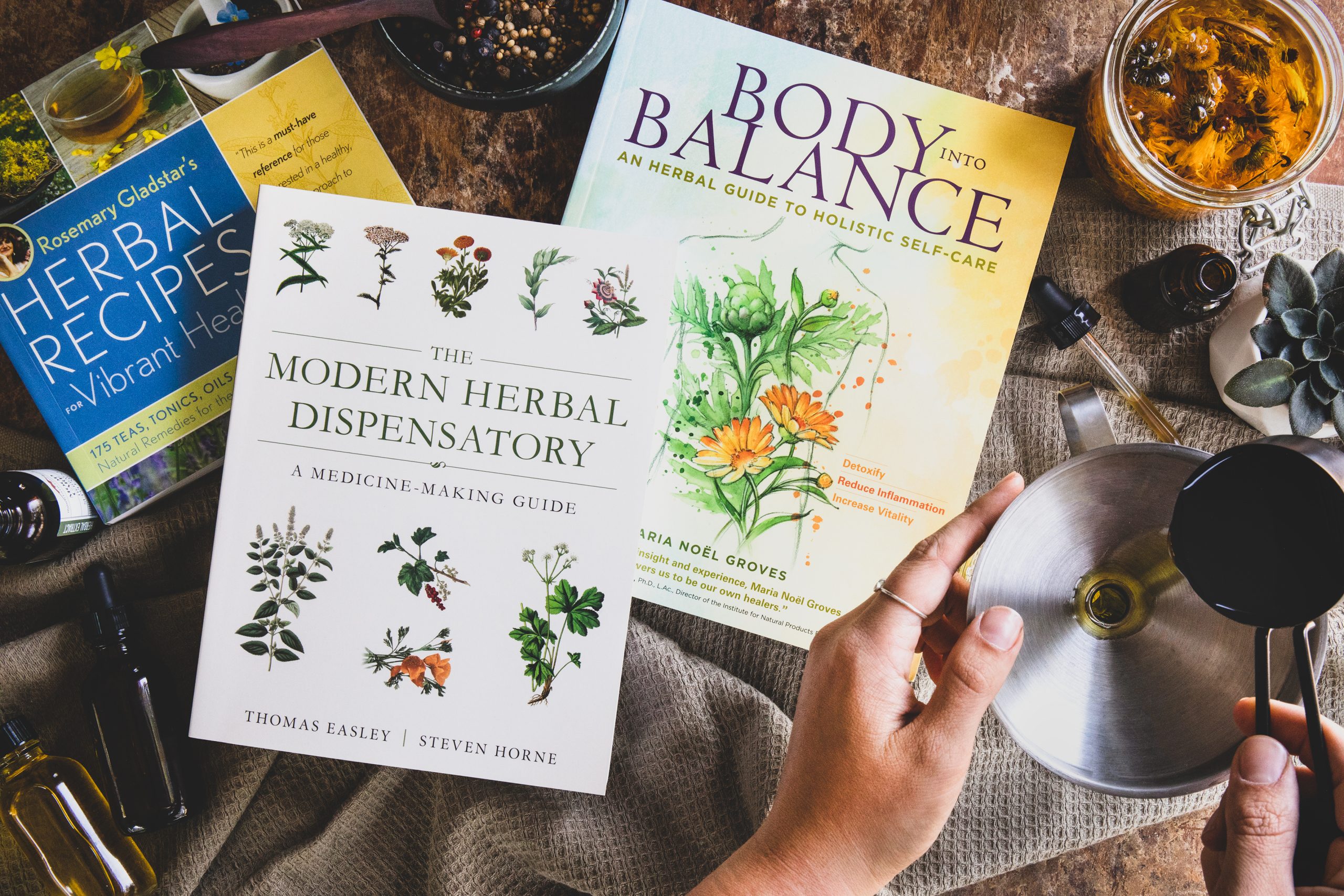 Three favorite books on herbalism with hands working on a recipe. 