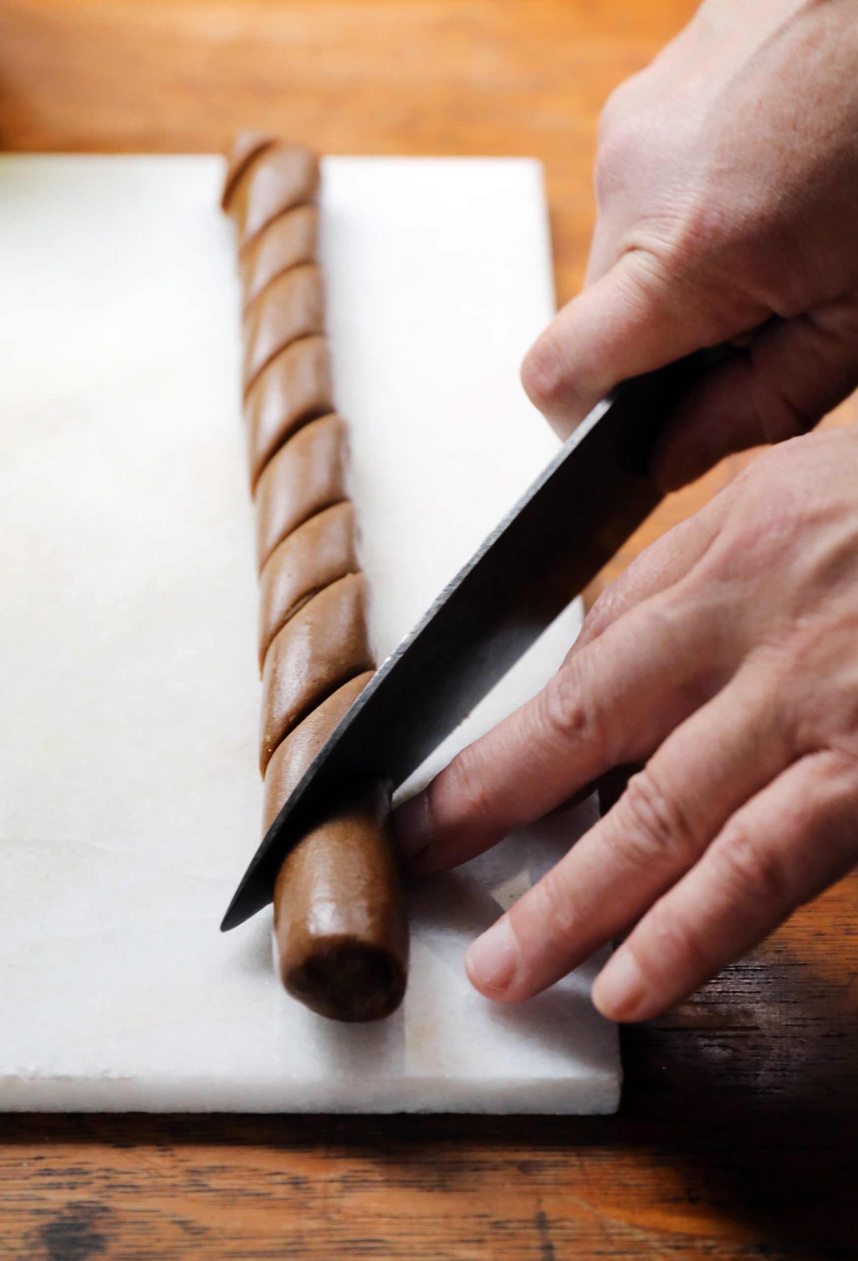 Roll of gingerbread peppernut dough being cut diagonally into diamonds on white cutting board.