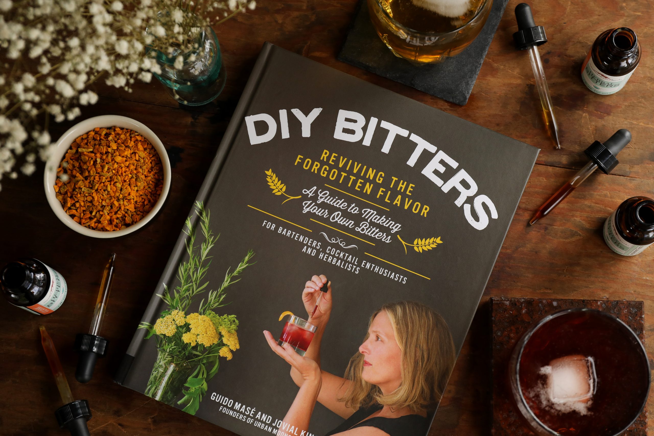 Hardback copy of book DIY Bitters: Reviving the Forgotten Flavor, by Guido Mas and Jovial King on a table surrounded by bottles of bitters, a cocktail made with bitters, and pipettes for measuring digestive bitters.