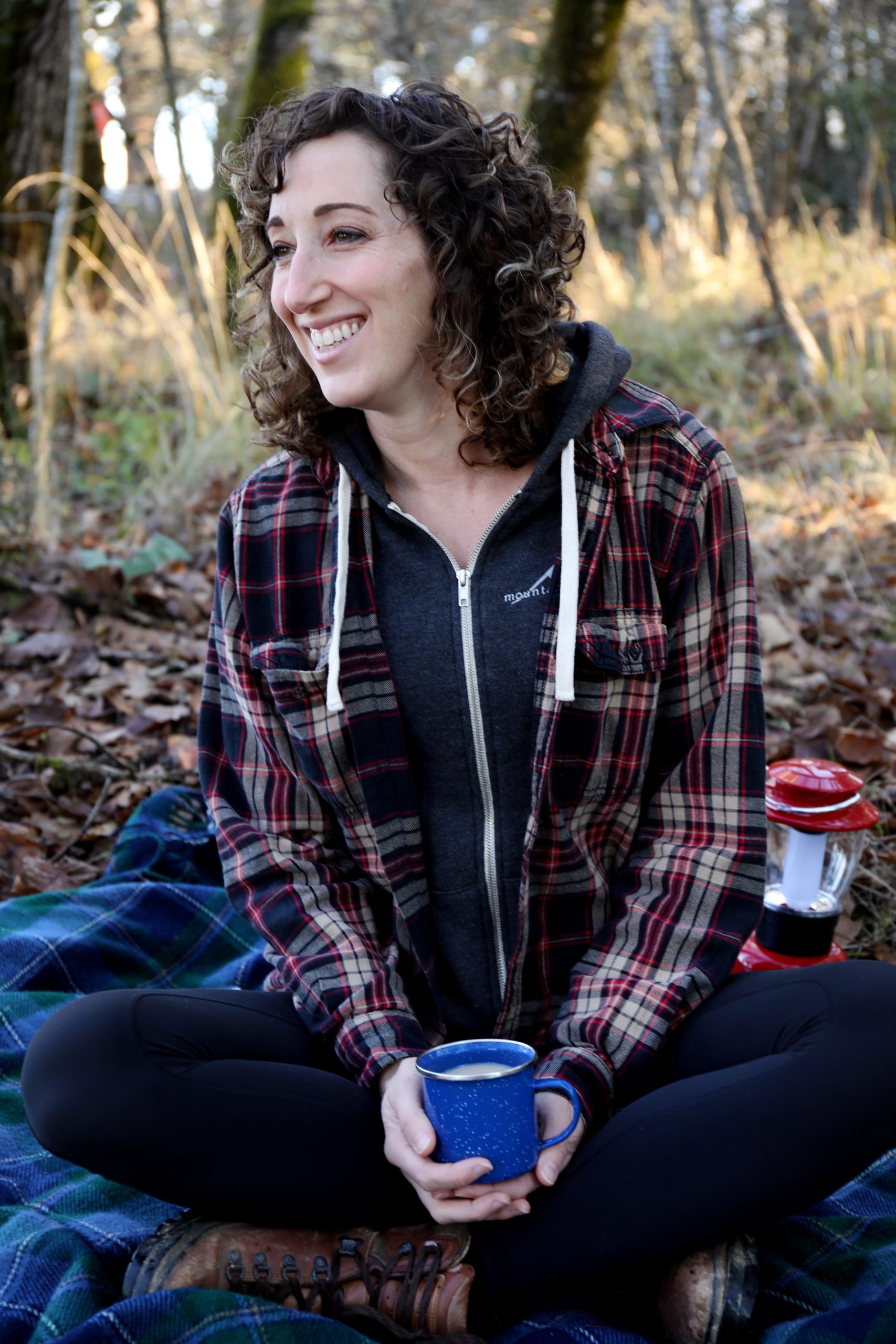 Young woman in a flannel shirt, smiling, while holding a warm beverage in a natural setting.