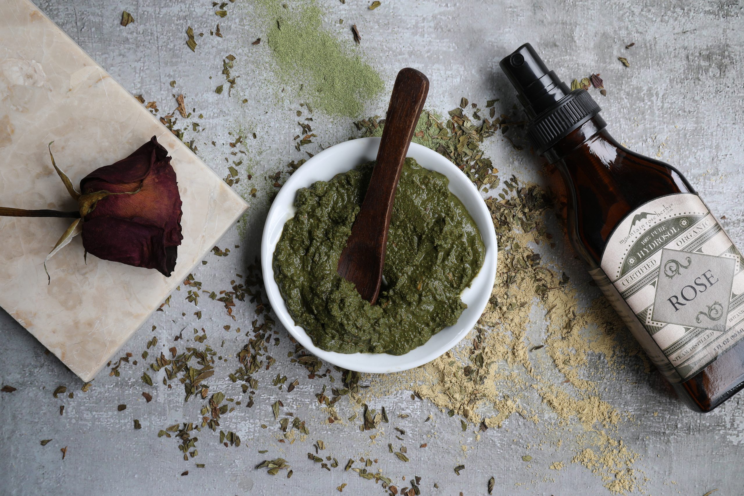 White bowl filled with green paste with displayed botanicals and a bottle of hydrosol. 
