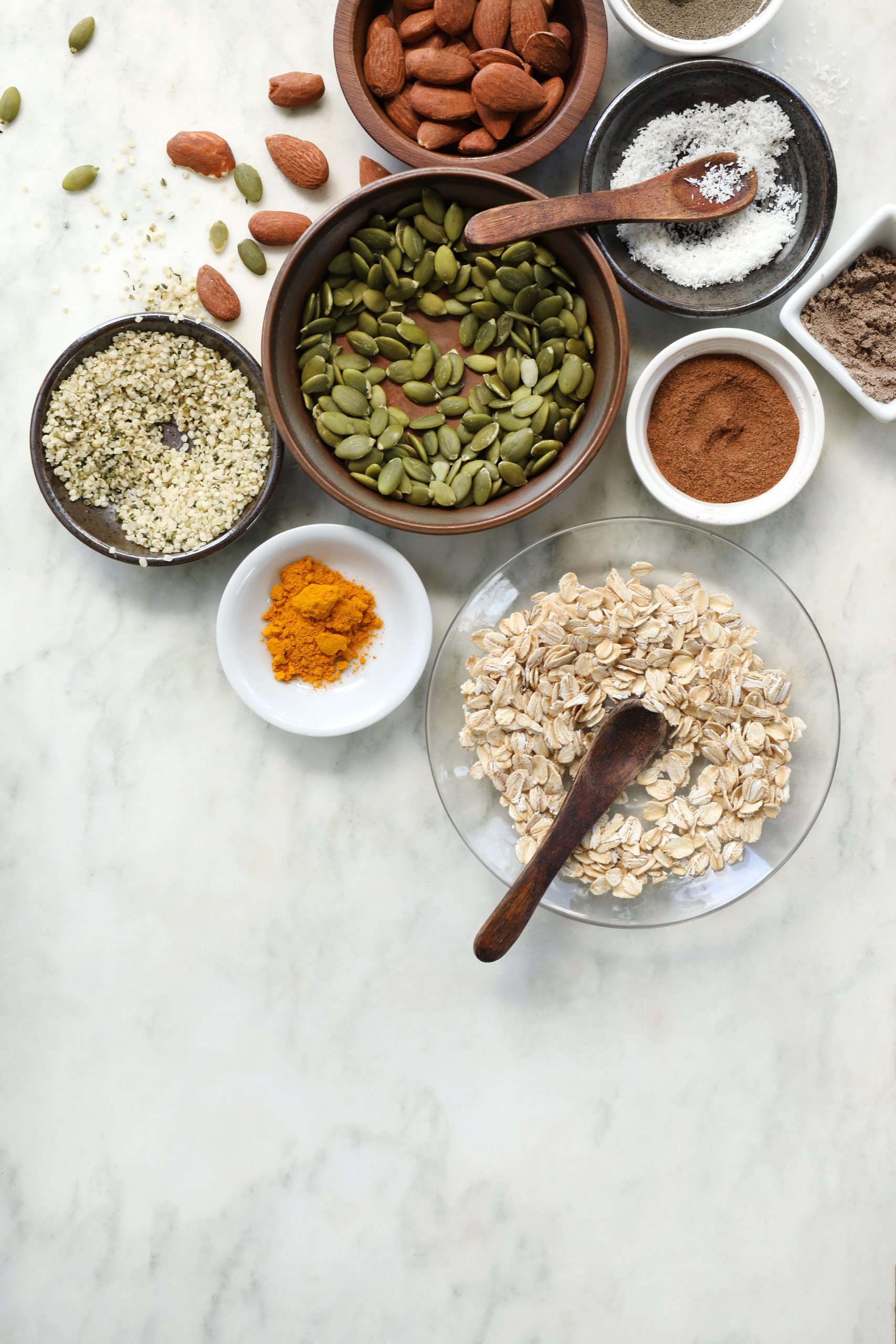 Ingredients are laid out to prepare a homemade granola featuring pepitas, almonds, whole oats, hemp seeds, and coconut flakes. Spices include turmeric, cardamom, cinnamon, and vanilla extract.