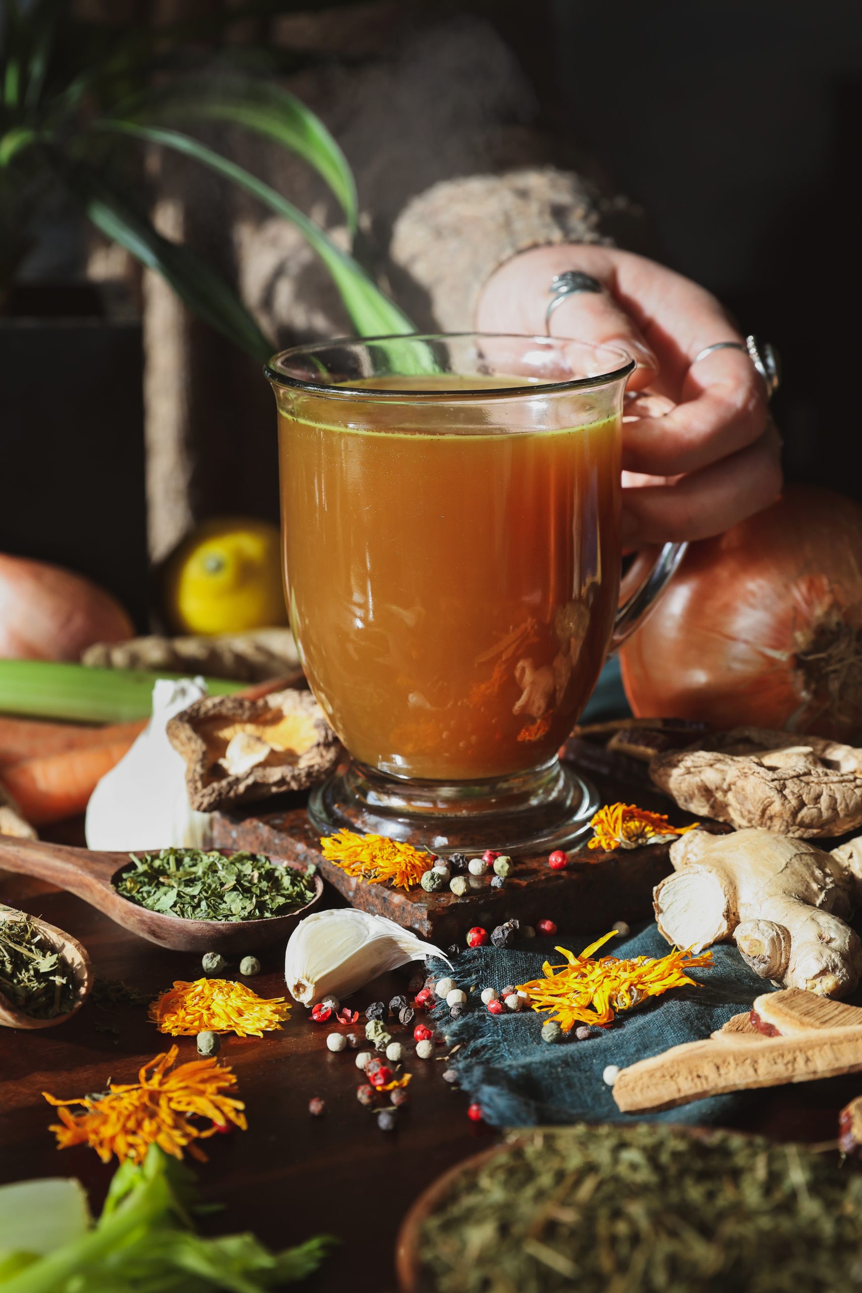 A mug of warm herbal broth with a hand reaching for the mug. The mug is surrounded by the ingredients of the herbal broth.