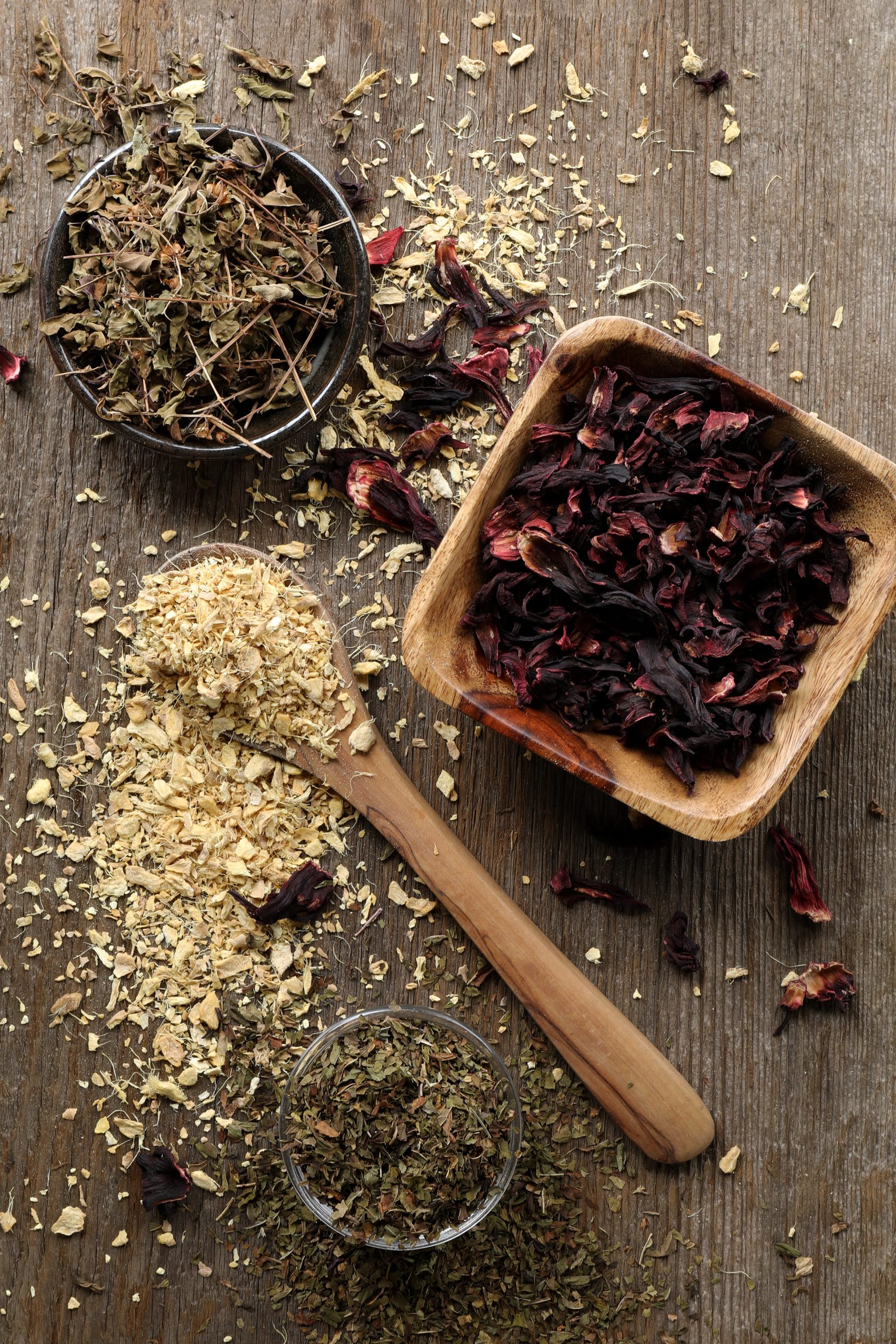 Herbs arranged on a rustic table top. Holy basil, dried ginger root, hibiscus flowers, and yerba mate tea in bowls and wooden spoons.