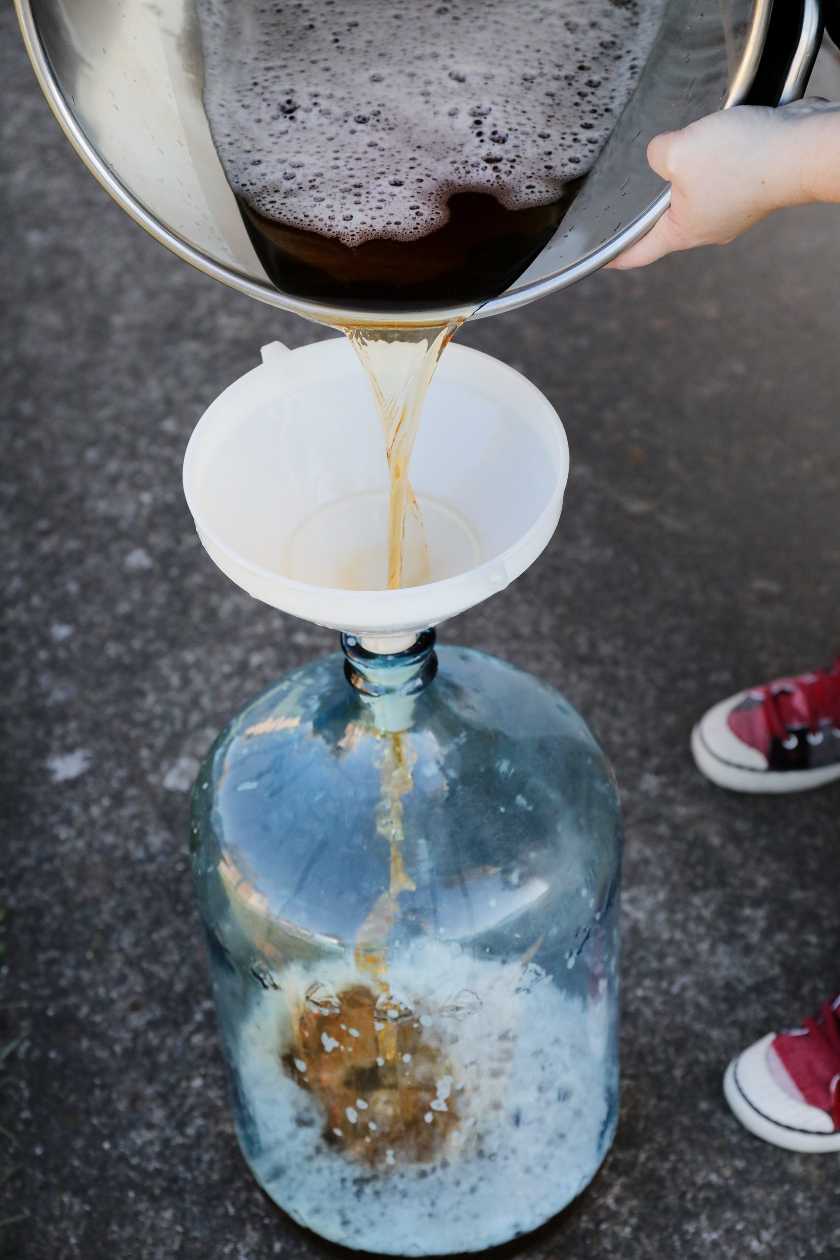Hands pouring colorful liquid through funnel into glass carboy. 