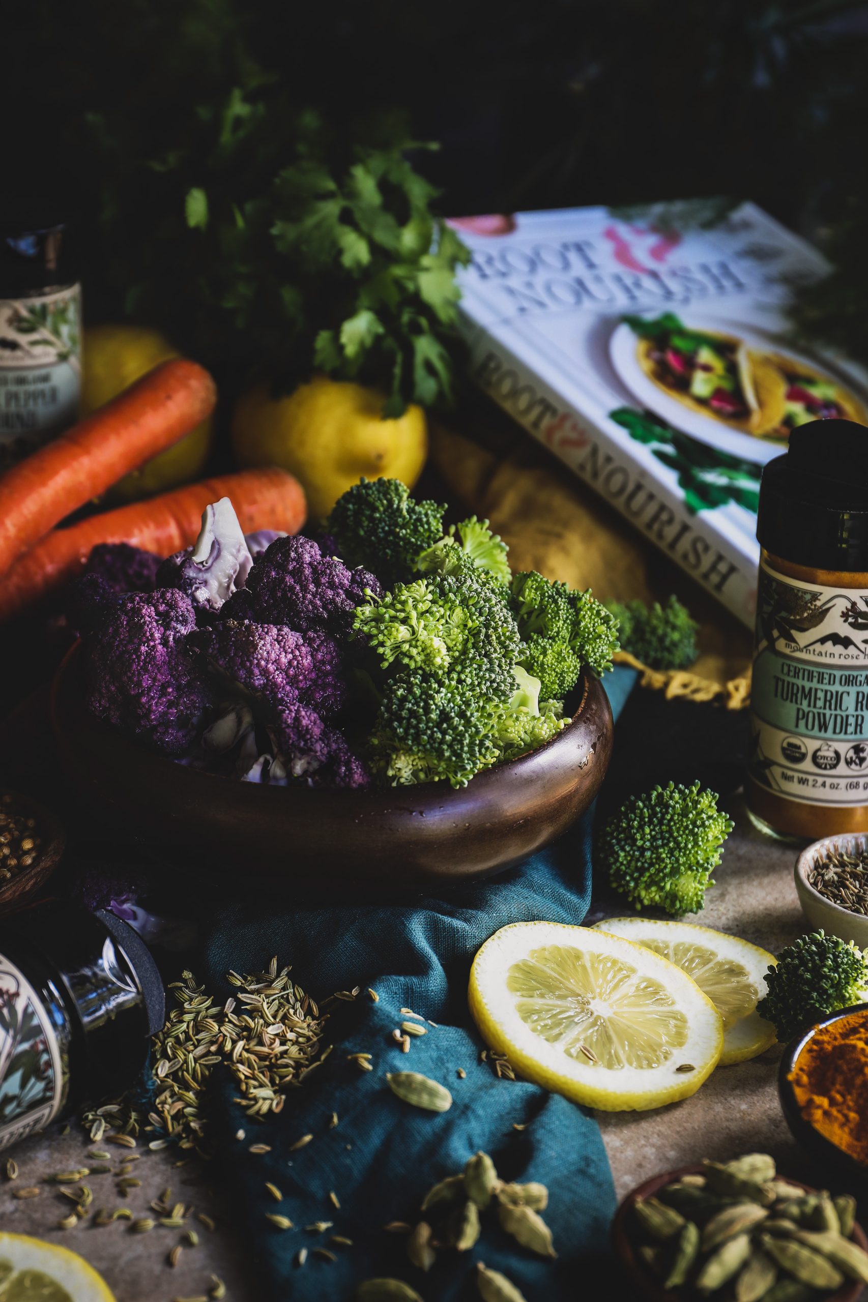 Wooden bowl of purple cauliflower, green broccoli surrounded by fresh spices and lemon slices.  Root and Nourish Cookbook in the background. 