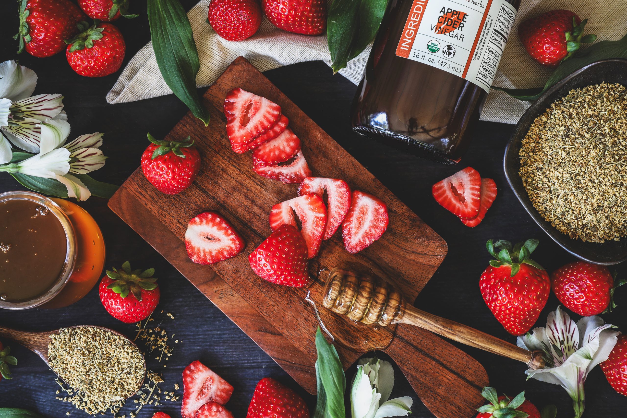 Fresh cut strawberries sit on cutting board with honey, apple cider vinegar, and elderflowers nearby