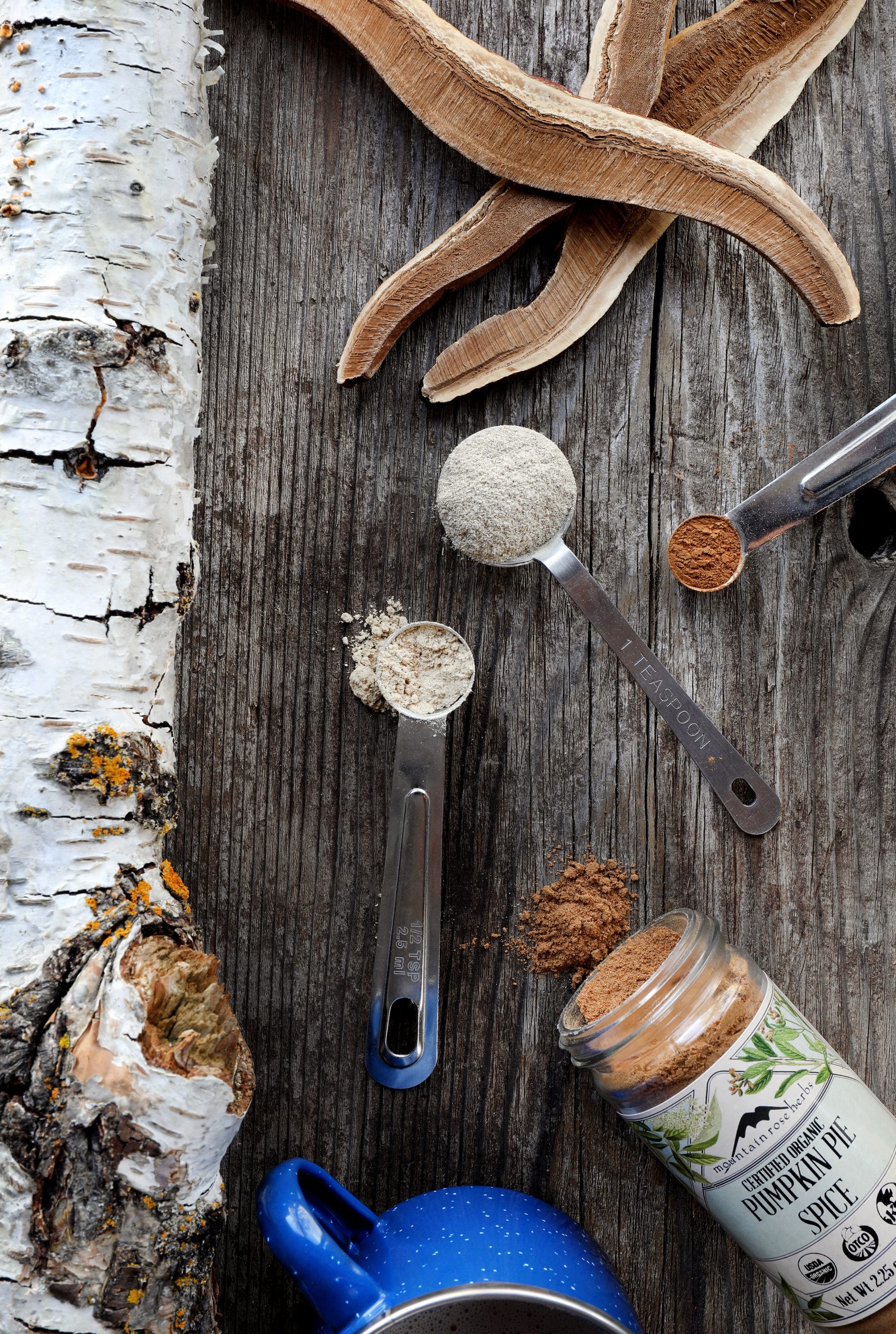Spoons of colorful powder, reishi mushroom slices and a bottle of pumpkin spice spilling into rustic board.