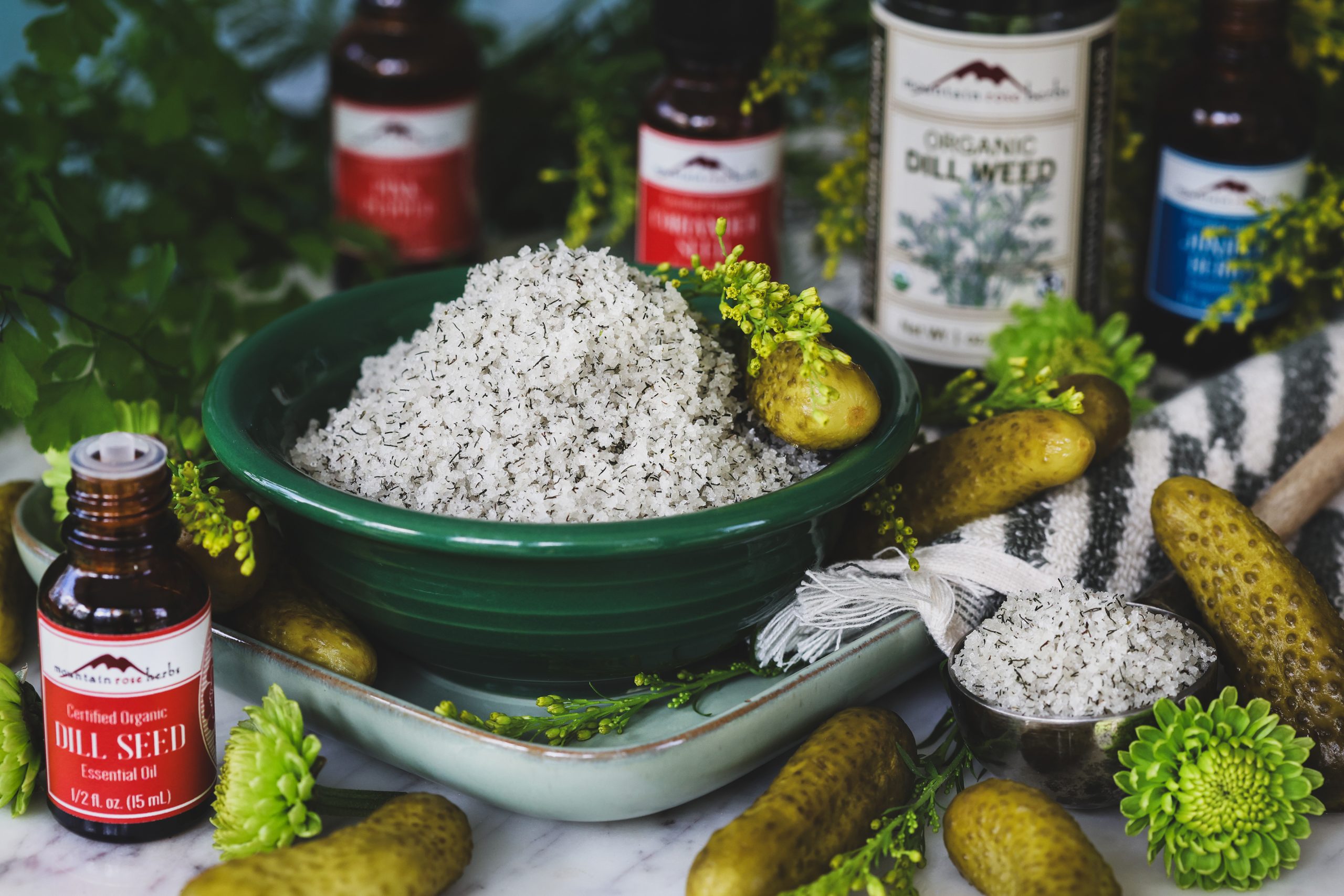 A bowl filled with pickle bath salts sits surrounded by pickles sits on a counter top