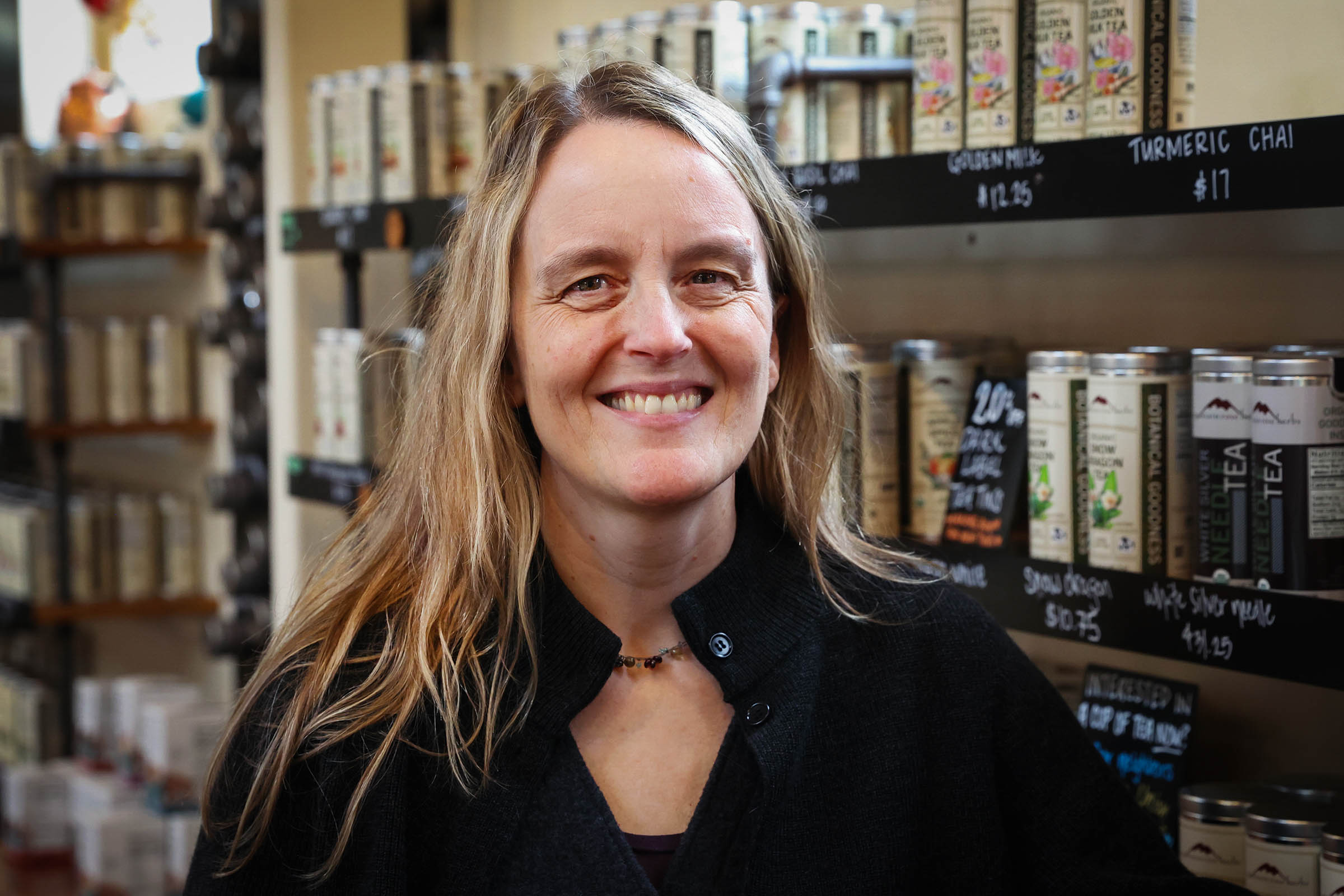 Cameron Stearns, CEO/CFO of Mountain Rose Herbs, poses in front of a wall of tea tins in the Mountain Rose Herbs Mercantile. She is wearing a black sweater and has light brown hair.