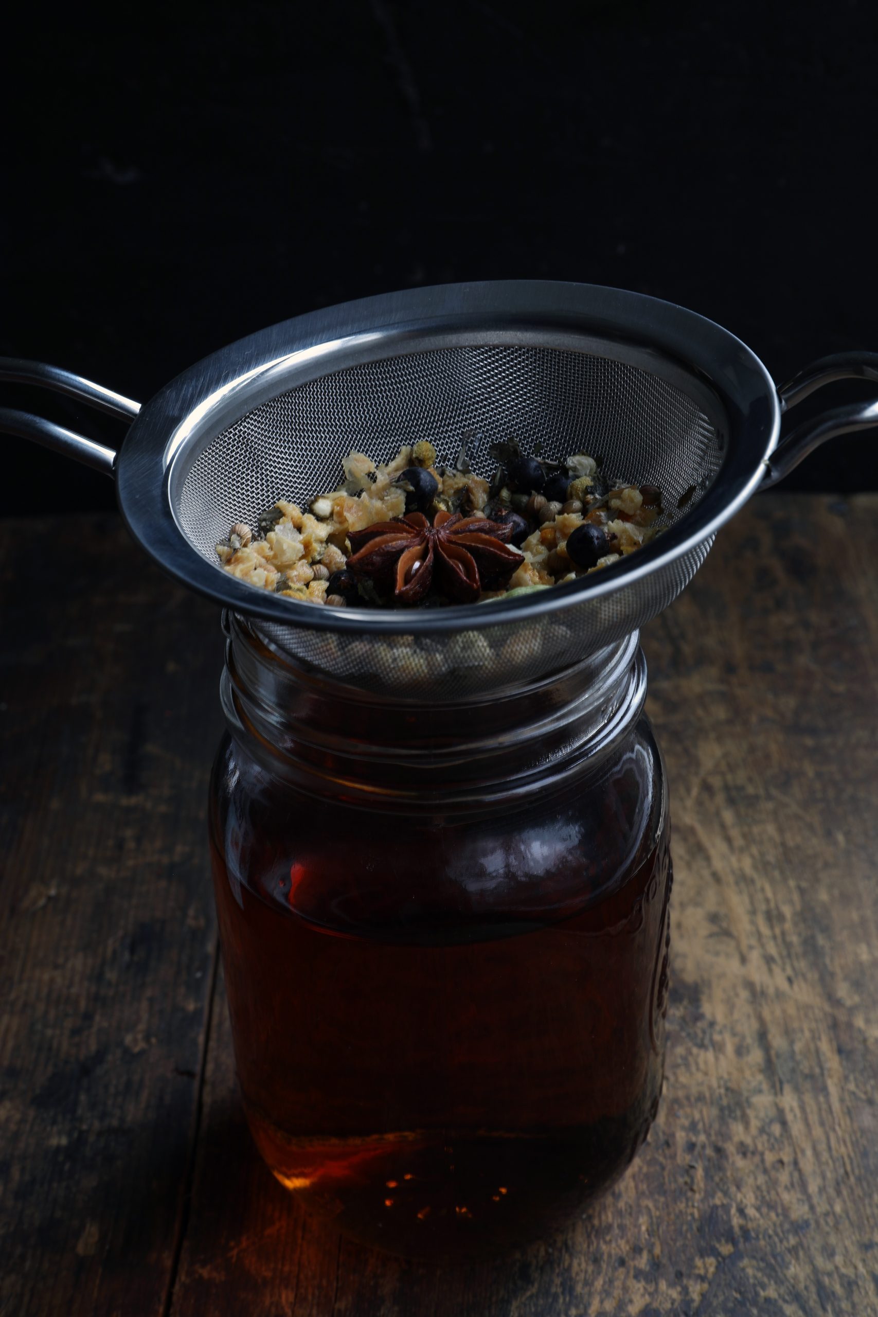 Strainer holding boiled herbs and spices over glass pantry jar with strained vermouth Strainer holding boiled herbs and spices over glass pantry jar with strained vermouth