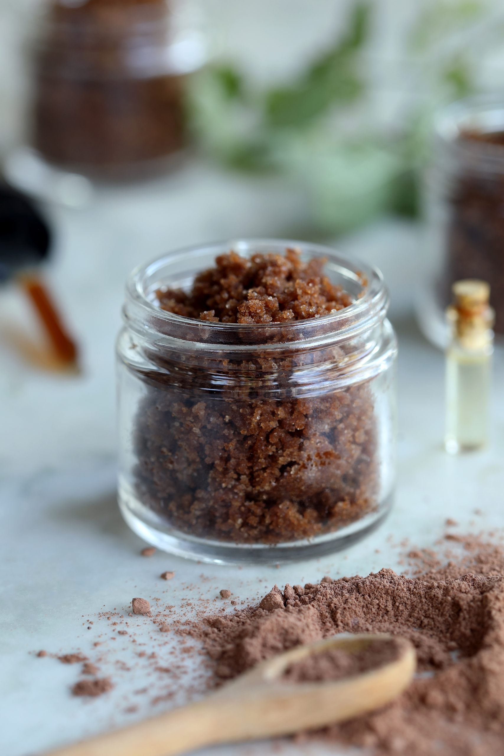 A vanilla body scrub using brown sugar, cacao powder, sunflower oil, and vanilla extract is stored in a clear glass salve jar and used for exfoliating and nourishing the skin.