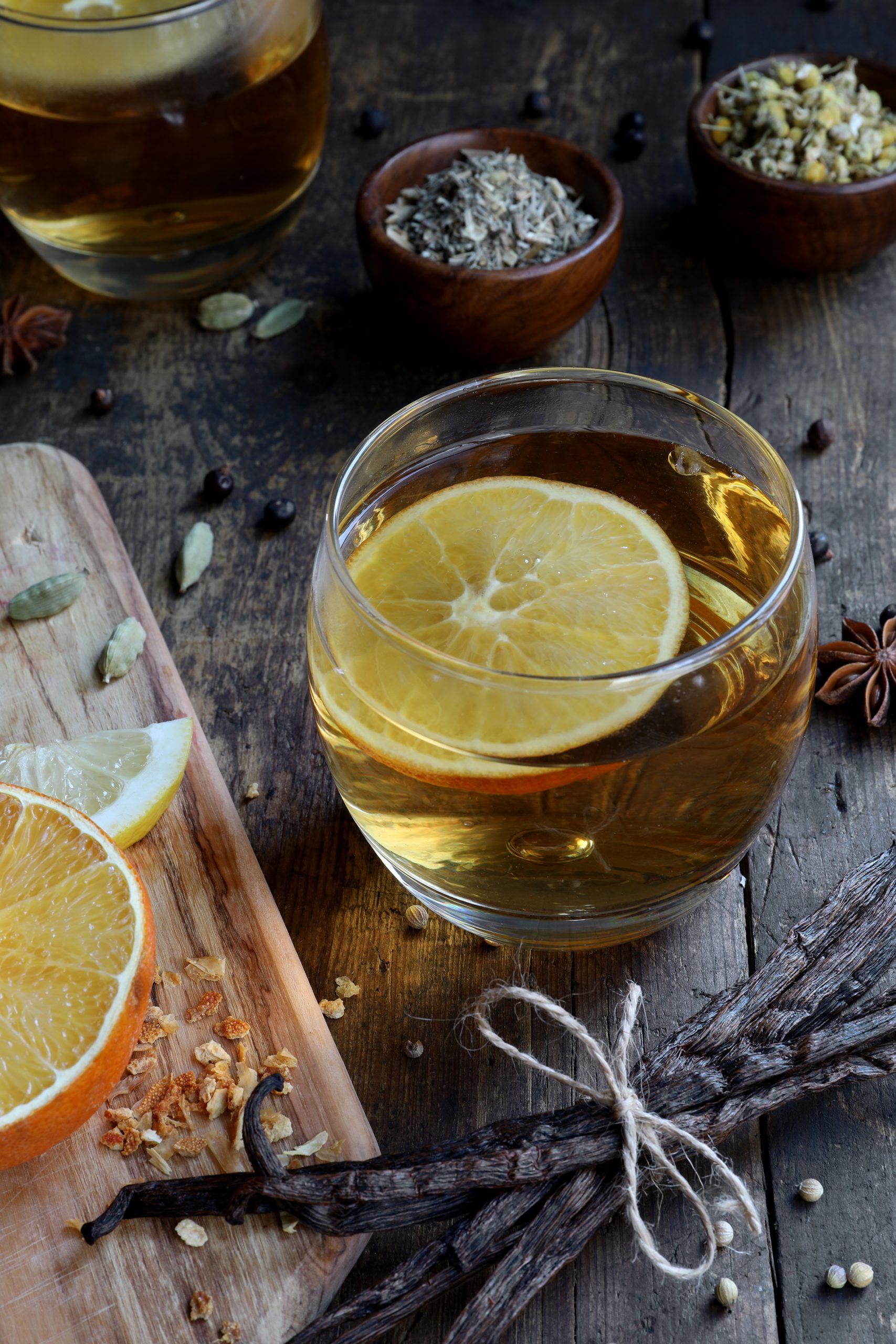 Glass cup filled with golden liquor, with an orange slice and various loose botanicals surrounding it, on a rustic table. 