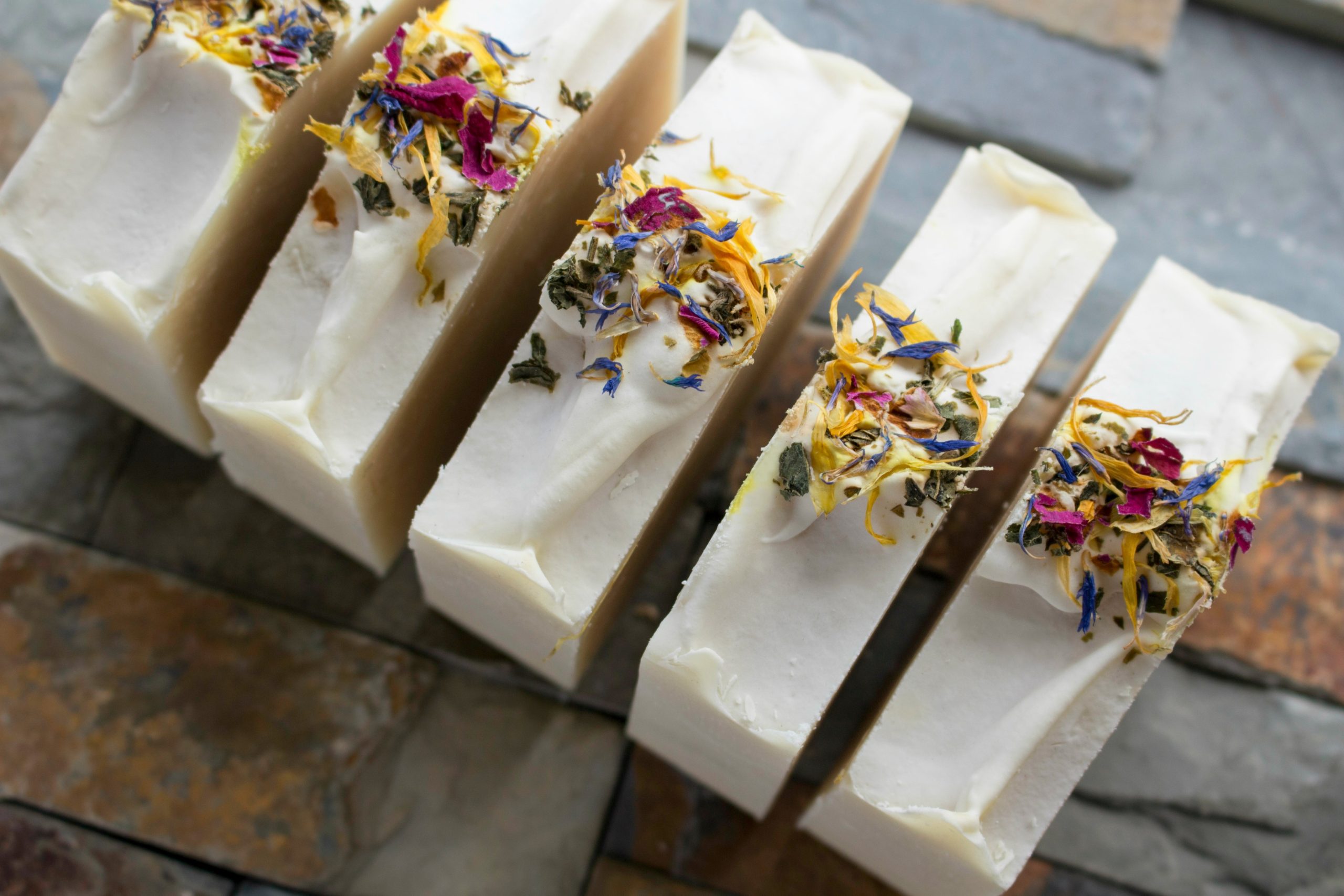 White bars of soap sprinkled with colorful flower petals on a cool-colored natural stones. 