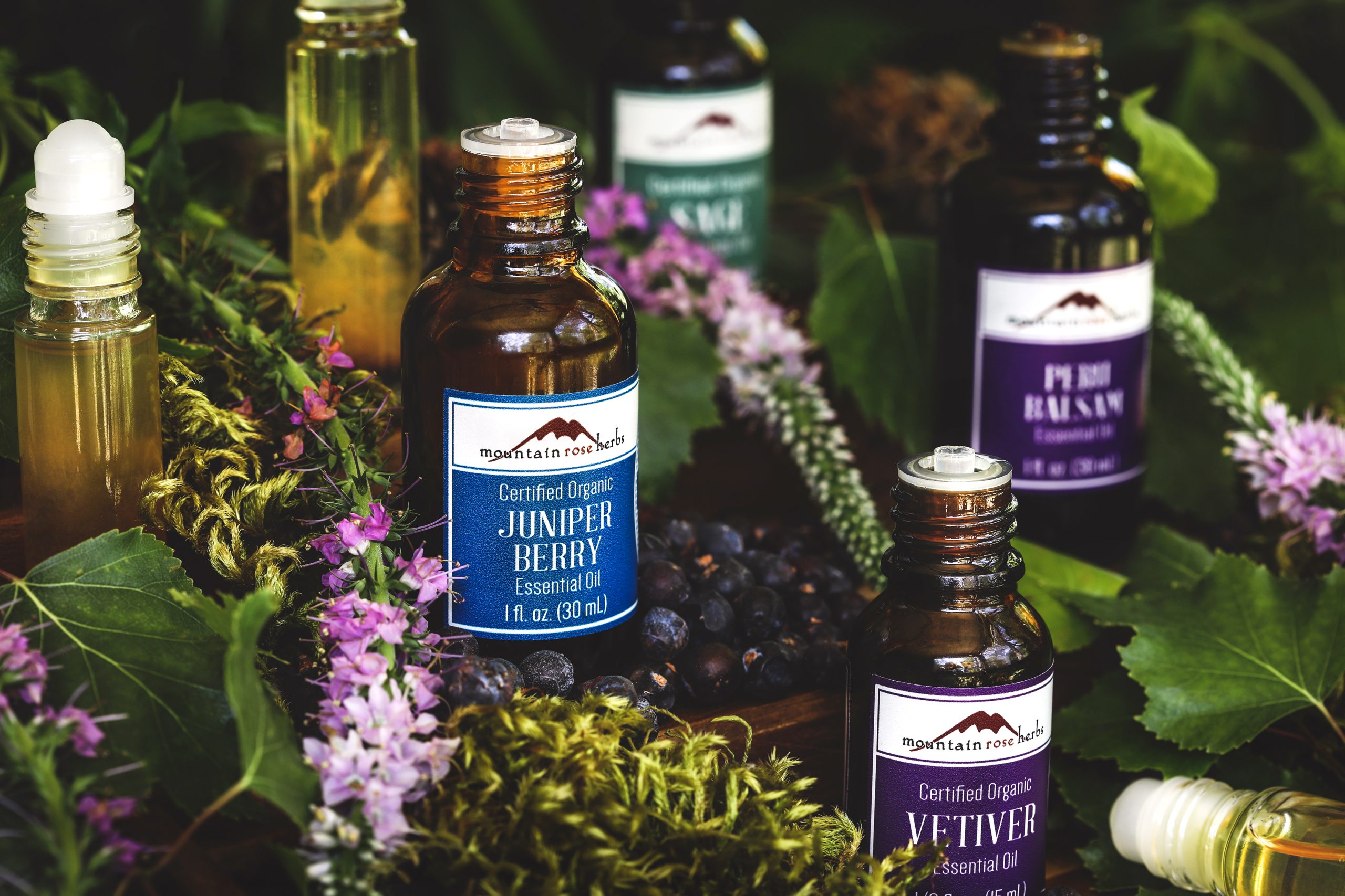 Bottles of essential oils used in the earth goddess lotion recipe surrounded by moss, fresh flowers, and spring green leaves. 