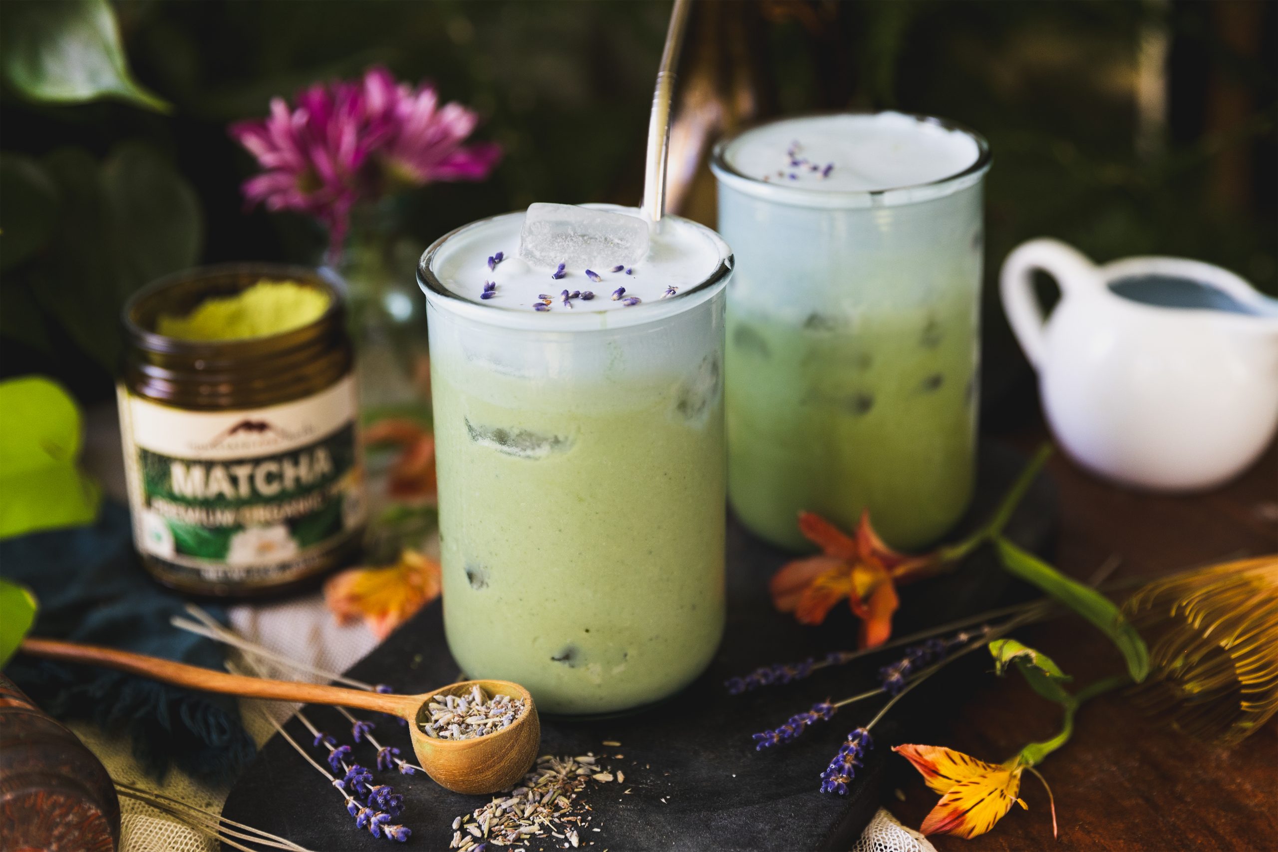 Two glasses of lavender matcha latte sit next to orange flowers, some lavender, and a jar of matcha powder.