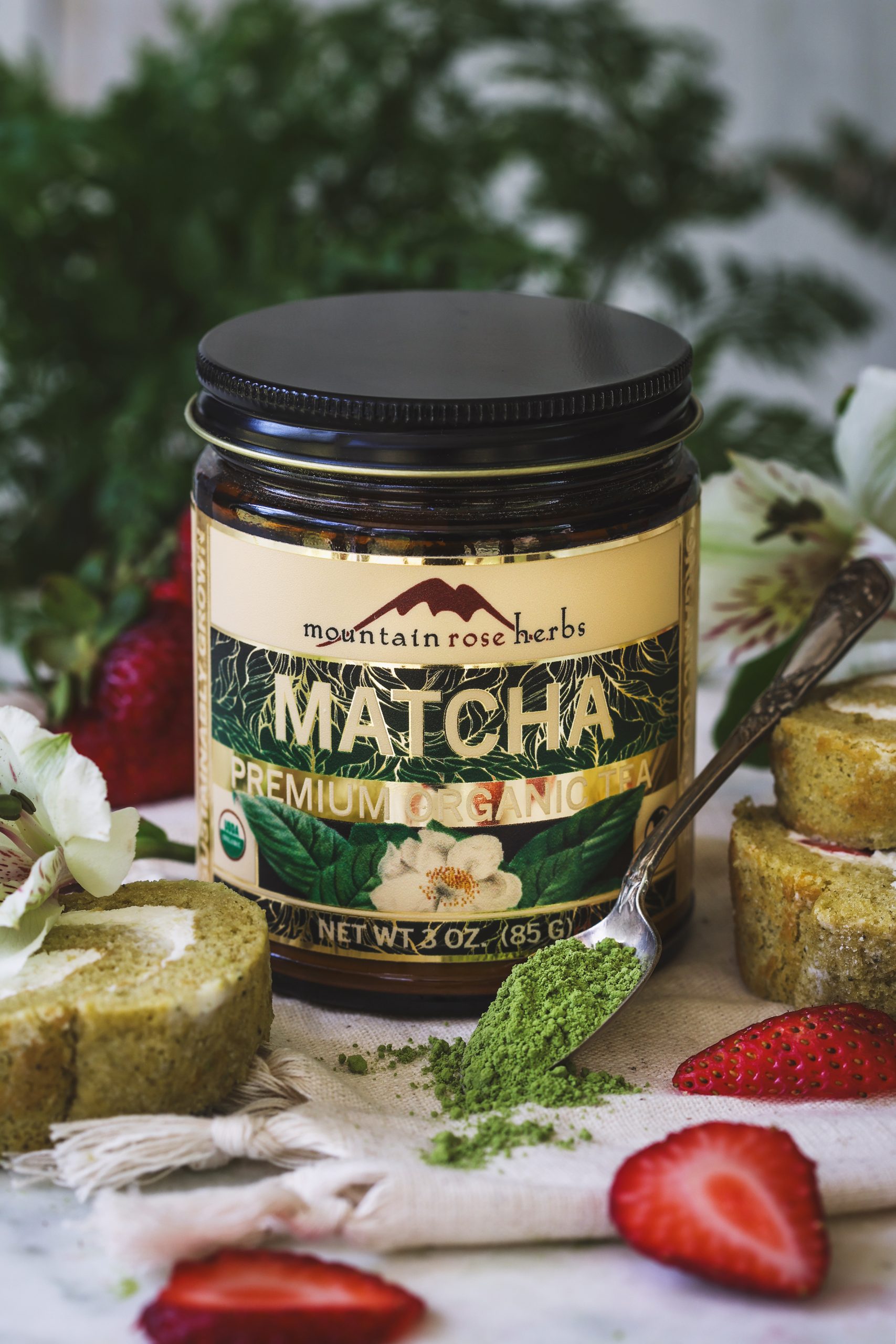Jar of Mountain Rose Herbs' premium matcha tea powder, surrounded by fresh strawberries and swiss roll slices.