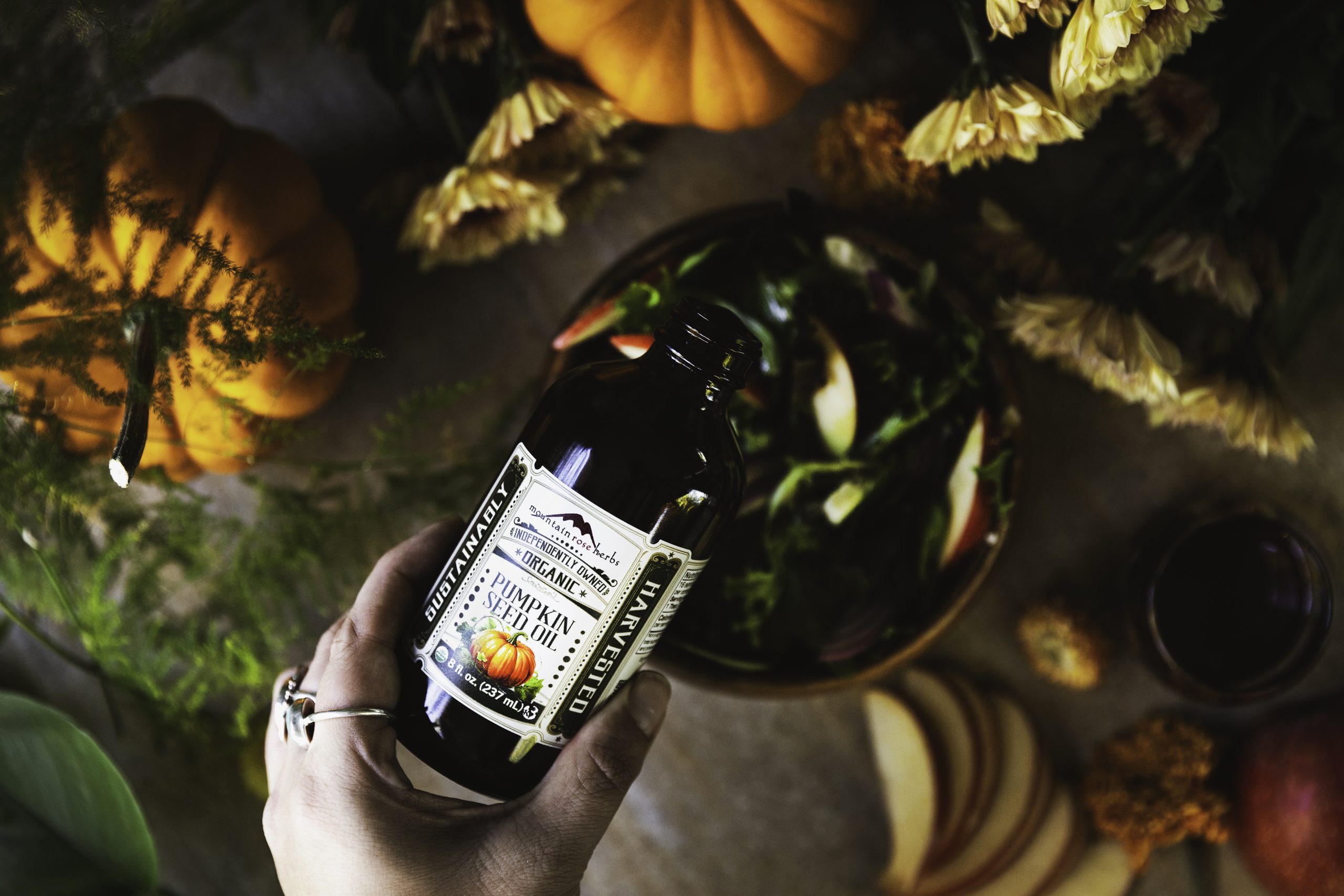 A bottle of Mountain Rose Herbs Pumpkin Seed Oil is being poured from a hand into a bowl of salad surrounded by fall items like small pumpkins and orange flowers.