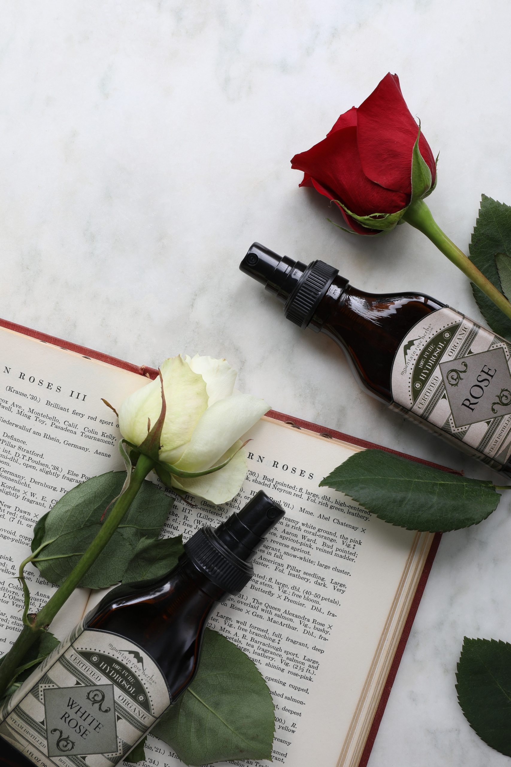 Glass bottles of white rose and rose hydrosols laying out near fresh roses and a book