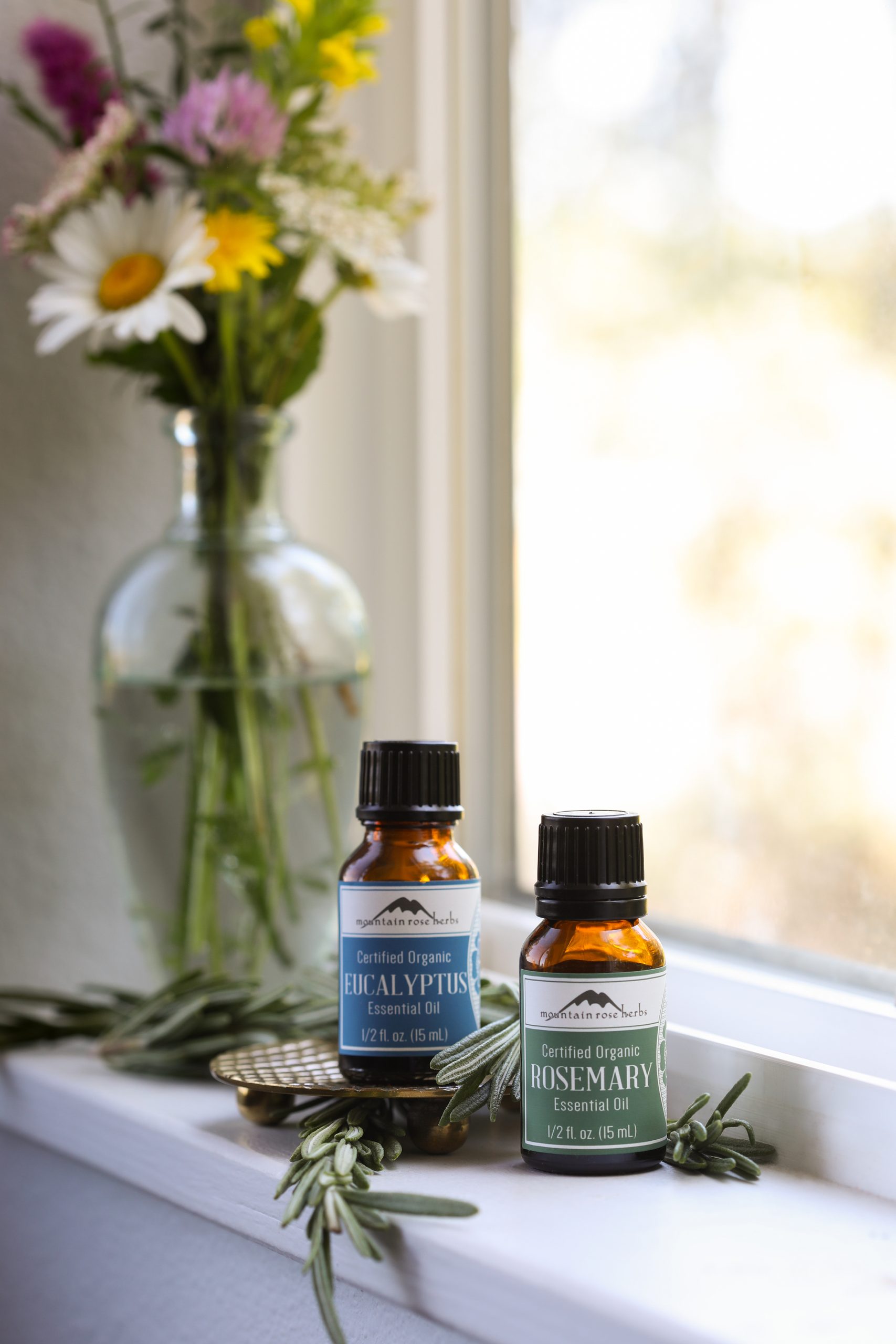 Bottles of eucalyptus and rosemary essential oils on a windowsill with fresh rosemary.