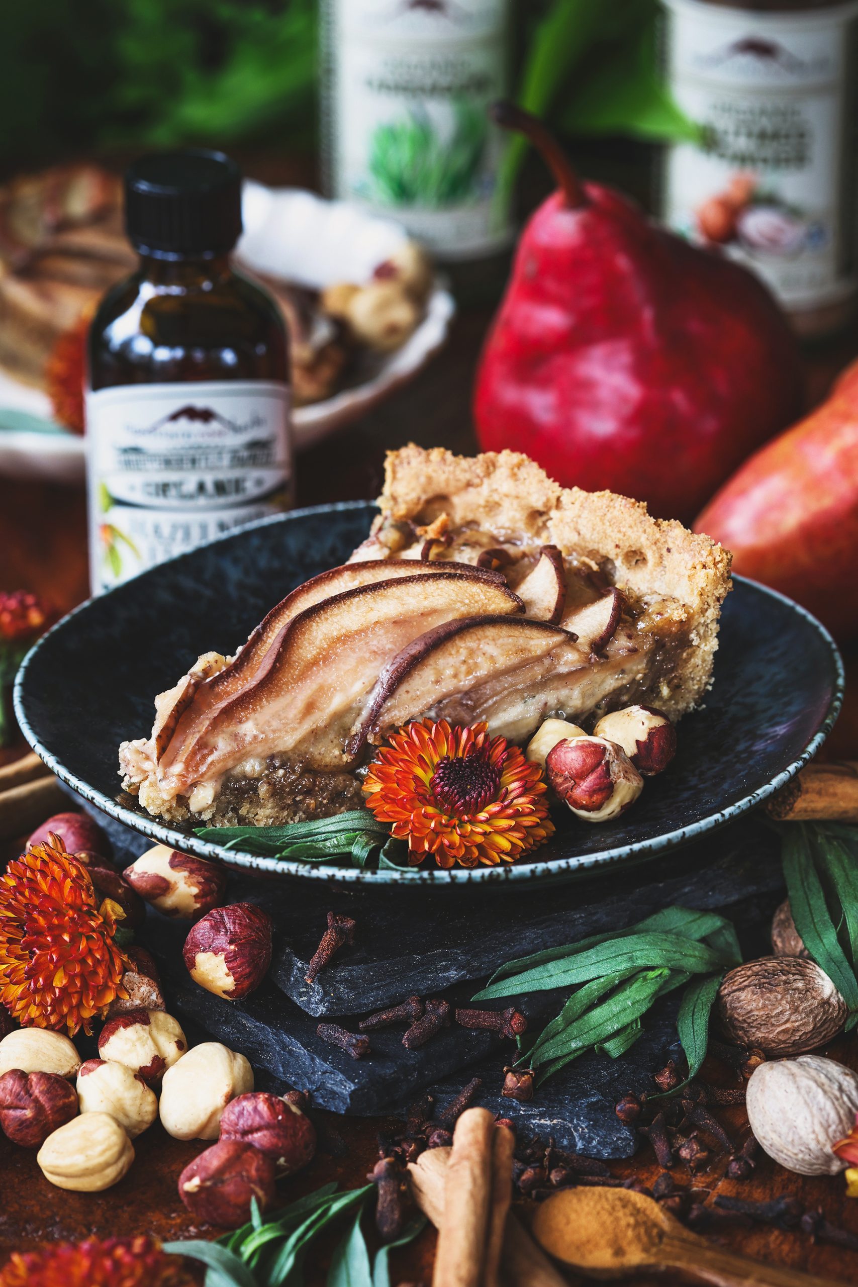 A slice of pear hazelnut tart on a plate with fresh tarragon and hazelnuts