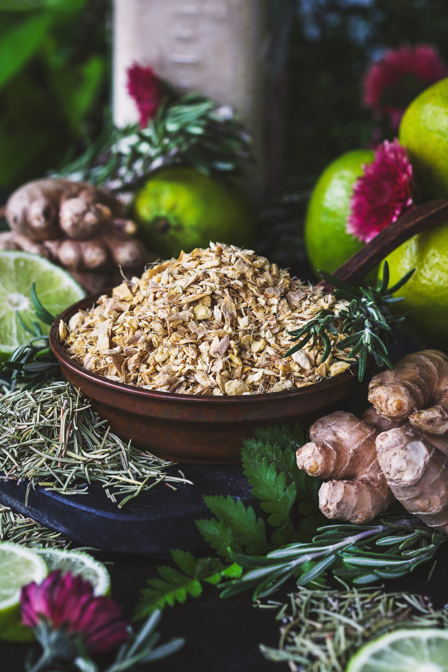 A close up photo of dried ginger root, and rosemary with fresh limes and ginger root in the background