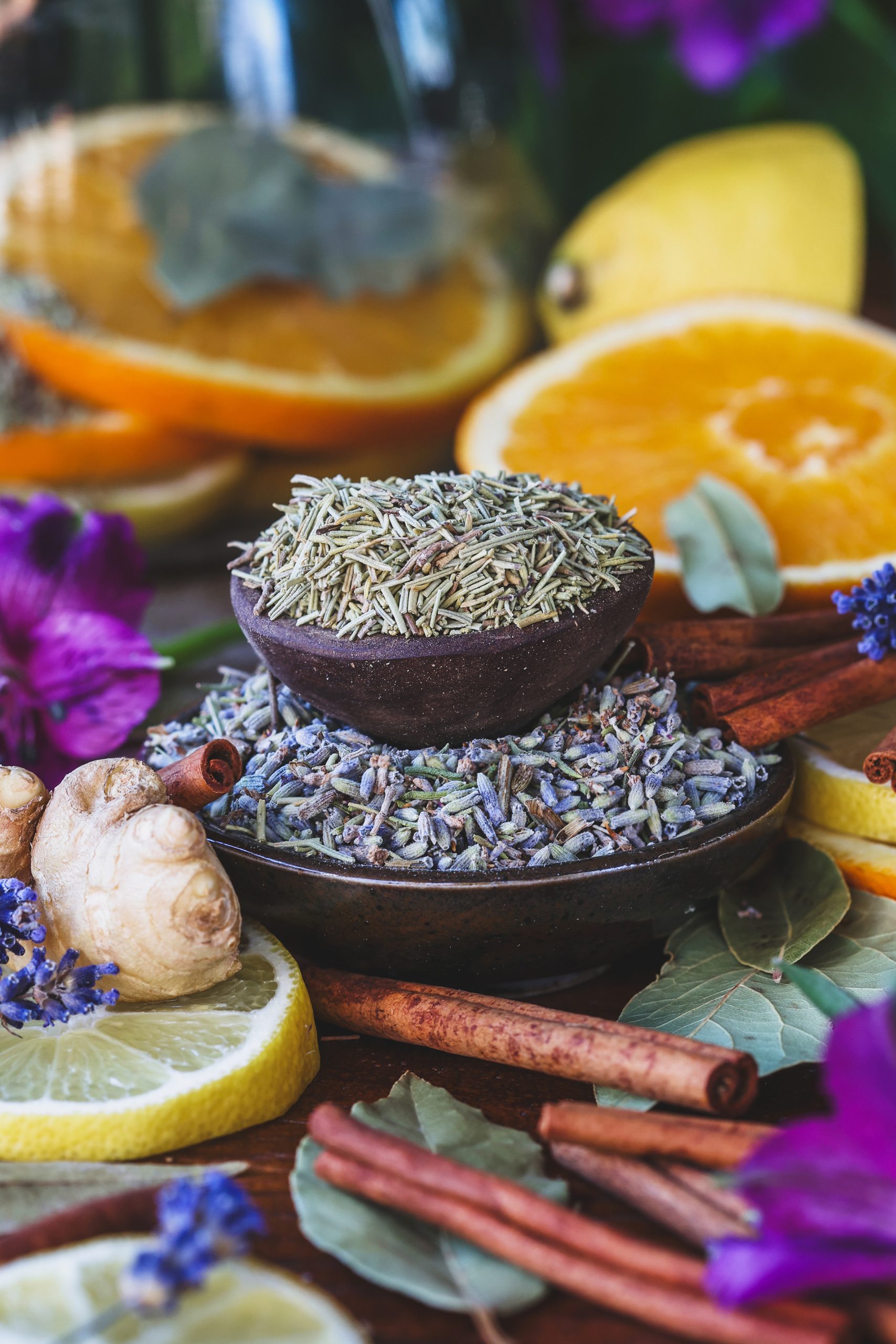 Two small bowls of dried lavender and rosemary with bay leaves, cinnamon sticks, fresh ginger, and citrus nearby