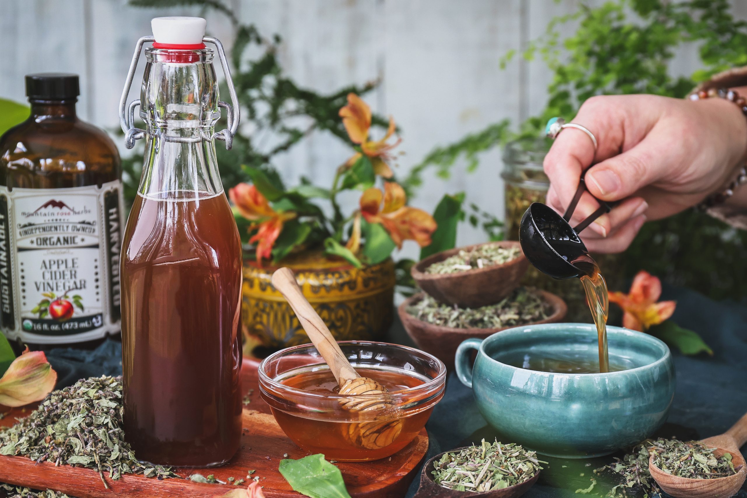A bottle of oxymel with honey and dried herbs around it with a cup nearby